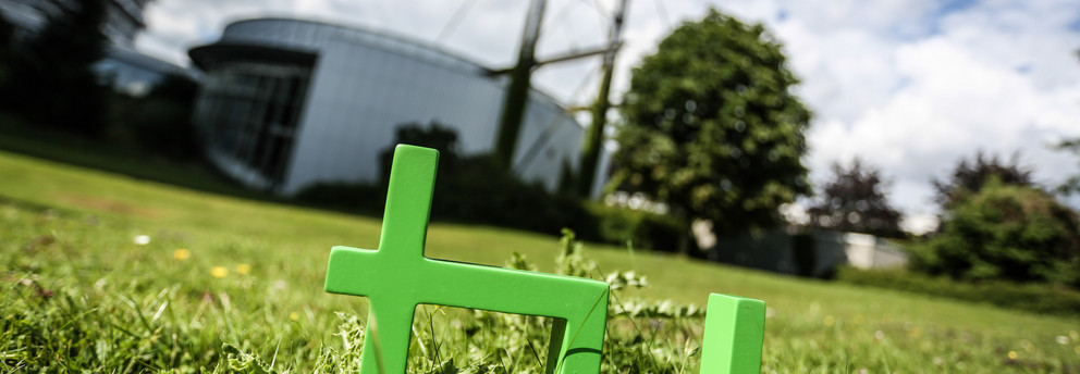 Im Vordergrund steht das TU Logo im Gras, im Hintergrund das Audimax.